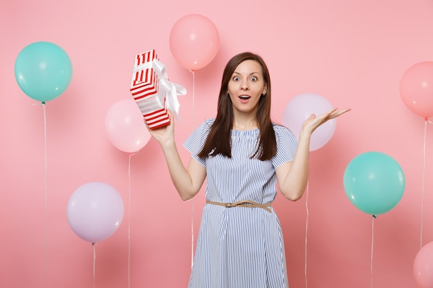 Retrato de mujer joven sorprendida en vestido azul extendiendo las manos sosteniendo la caja roja con regalo presente sobre fondo rosa con coloridos globos aerostáticos. Fiesta de cumpleaños, concepto de emociones sinceras de personas.