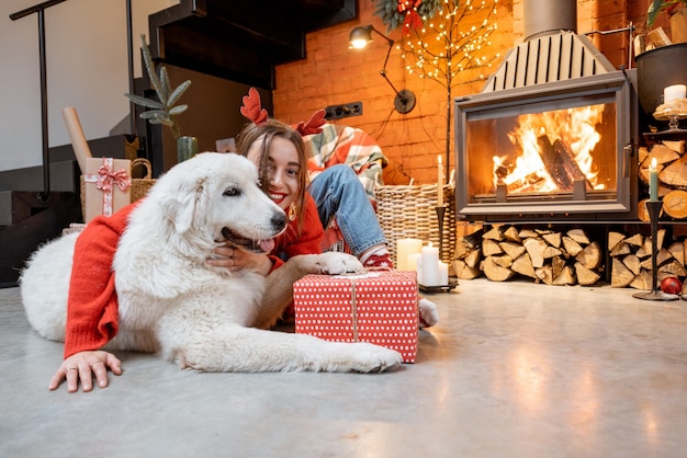 Retrato de una mujer joven con su lindo perro blanco desembalaje de cajas de regalo junto a una chimenea durante unas felices vacaciones de Año Nuevo en casa