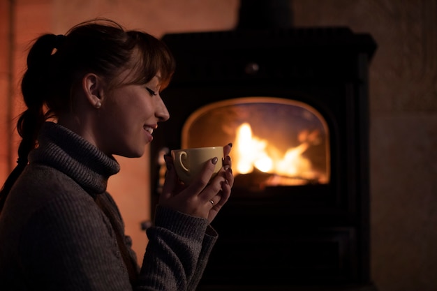 Retrato de mujer joven en un suéter caliente bebiendo bebida caliente de taza junto a la chimenea