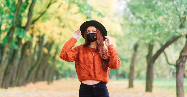 Retrato de mujer joven en suéter rojo tejido, sombrero en una máscara médica con estilo negro en el parque amarillo de otoño