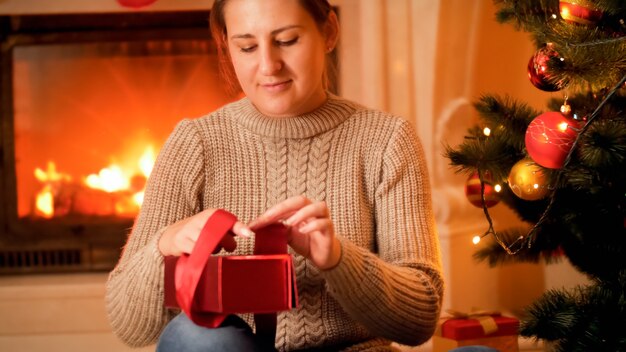 Retrato de mujer joven en suéter sentado junto a la quema de firepalce y empacando regalos y regalos de Navidad