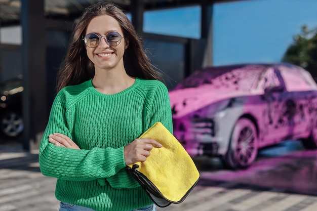 Retrato de mujer joven en suéter verde con tela de microfibra amarilla y camión cubierto con espuma de jabón rosa detrás