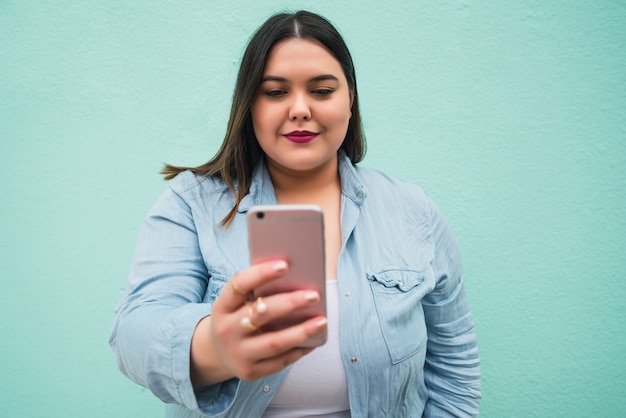 Retrato de mujer joven de talla grande haciendo videollamada en su teléfono móvil al aire libre contra el azul claro