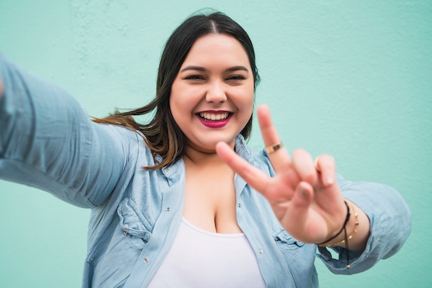 Retrato de mujer joven de talla grande tomando un selfie mientras está de pie contra la pared azul claro, al aire libre.