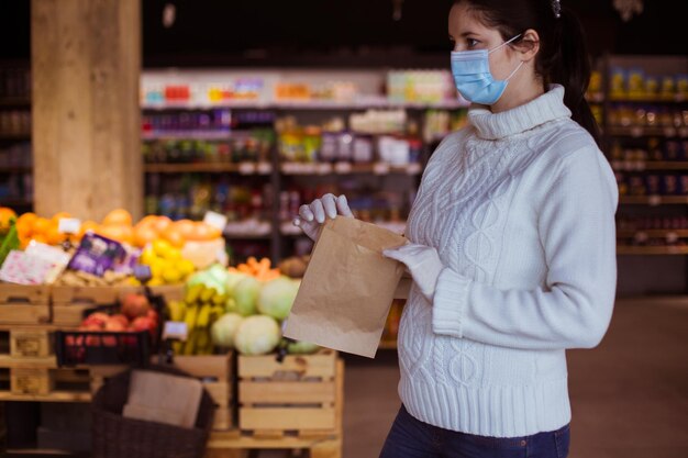 Retrato de mujer joven en la tienda de comestibles Asistente de tienda en guantes protectores y máscara médica posando con bolsa de papel con espacio de copia en sus manos