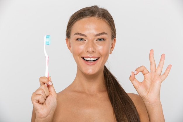 Retrato de una mujer joven en topless feliz aislada, sosteniendo el cepillo de dientes con una pasta de dientes, mostrando ok