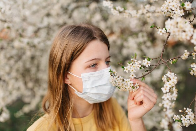 Foto retrato de mujer joven triste en mascarilla médica protectora con flores cerca de árbol floreciente en primavera. concepto de alergia de primavera