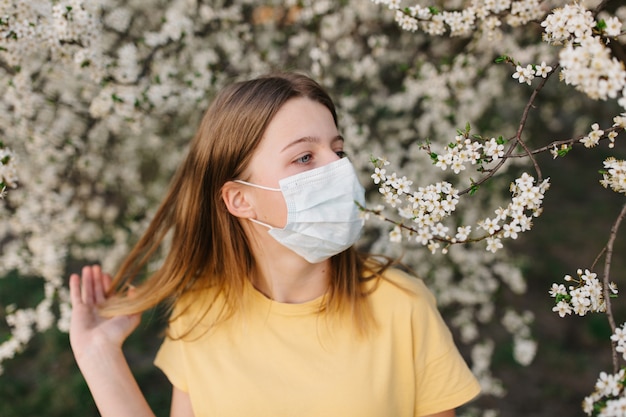 Retrato de mujer joven triste en mascarilla médica protectora con flores cerca de árbol floreciente en primavera. Concepto de alergia de primavera