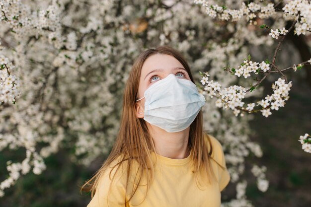 Retrato de mujer joven triste en mascarilla médica protectora con flores cerca de árbol floreciente en primavera. Concepto de alergia de primavera