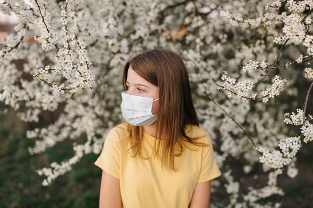 Retrato de mujer joven triste en mascarilla médica protectora con flores cerca de árbol floreciente en primavera. Concepto de alergia de primavera