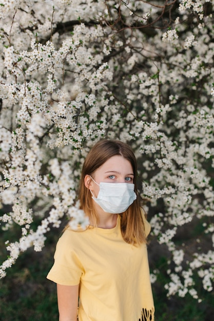 Retrato de mujer joven triste en mascarilla médica protectora con flores cerca de árbol floreciente en primavera.