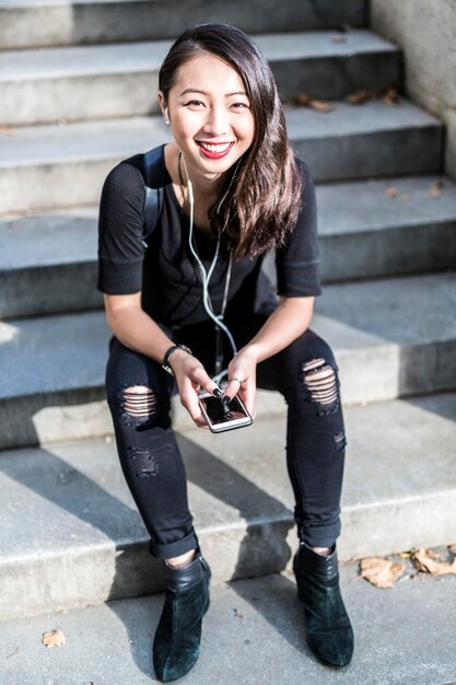 Foto retrato de mujer joven vestida de negro sentada en pasos escuchando música con auriculares y ph celular