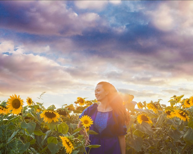 Retrato de una mujer joven con un vestido azul oscuro está de pie entre girasoles sobre un fondo de luz solar