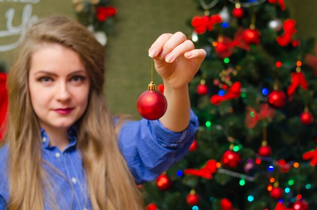 Retrato de una mujer con labios rojos brillantes, cabello largo rubio contra el árbol de año nuevo. Mujer joven en una camisa de hombre azul. sosteniendo una decoración navideña delante de él. Comiendo una pelota