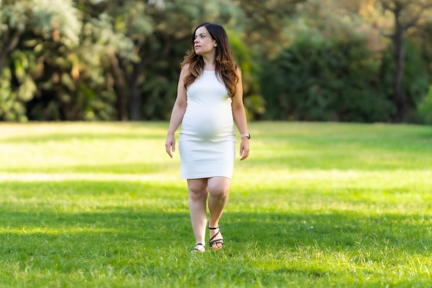 Retrato de una mujer latina embarazada adulta caminando sobre el césped de un parque con un vestido blanco de verano con una expresión distraída.