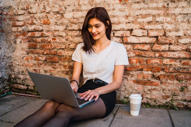 Retrato de mujer latina joven relajarse y usar la computadora portátil mientras está sentado al aire libre en la pared de ladrillo. Concepto urbano.