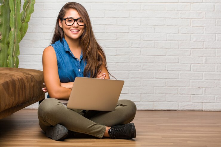 Foto retrato de una mujer latina joven sentada en el suelo cruzando los brazos, sonriendo y feliz, confiada y amigable. sosteniendo una laptop