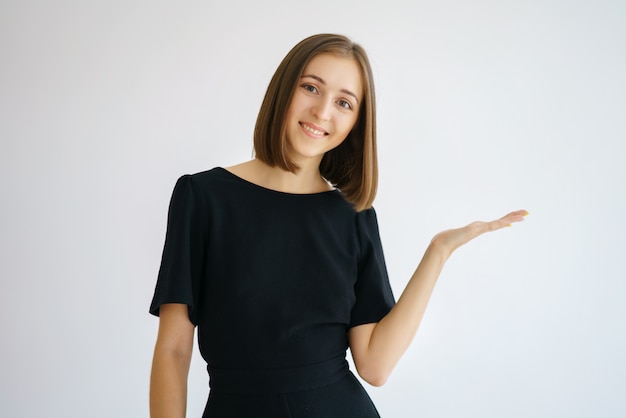 Retrato de una mujer linda feliz en un vestido negro sobre un fondo blanco sonriendo y mostrando un gesto con la mano a un lado, concepto