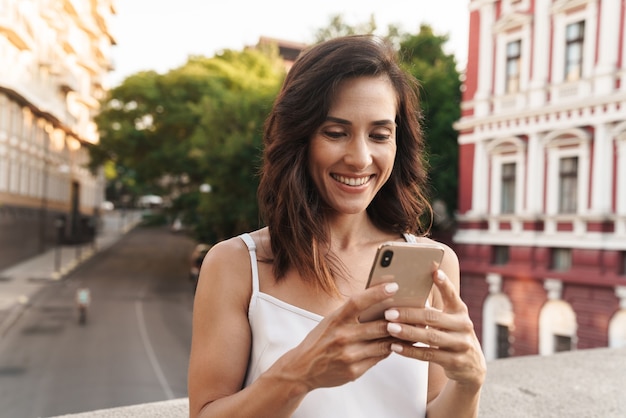Foto retrato de mujer linda sonriendo y escribiendo en el teléfono inteligente mientras está de pie en el puente con las calles de la ciudad