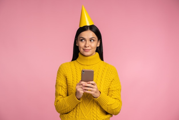 Retrato de mujer llena de alegría con sombrero de cono gracioso sosteniendo smartphone en sus manos y mirando a otro lado. Celebrando el concepto de vacaciones y tecnologías.