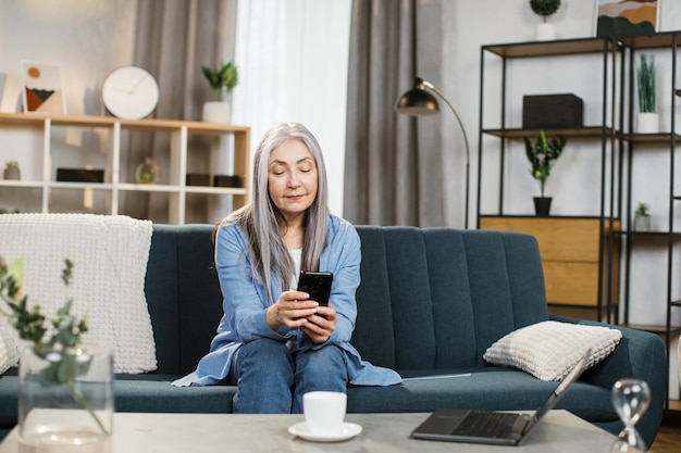 Retrato de una mujer madura alegre que usa un teléfono inteligente en casa para enviar mensajes o navegar por las redes sociales