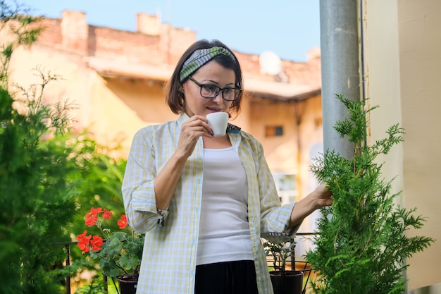 Retrato de mujer madura ama de casa, mujer con una taza de café en el balcón abierto decorado con plantas verdes