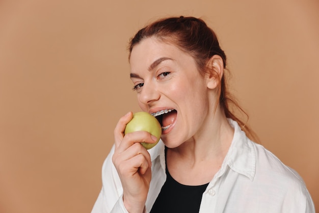 Retrato de una mujer madura con aparatos dentales comiendo una manzana verde sobre un fondo beige