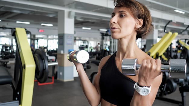 Retrato de mujer madura atlética está haciendo ejercicio de repeticiones para bíceps con pesas en las manos en el gimnasio. Ella está levantando pesas, copie el espacio. Concepto de entrenamiento y deporte.