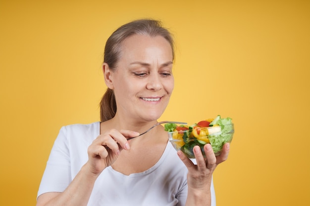 Retrato de una mujer madura contenta con una camisa blanca sosteniendo un plato de ensalada y un tenedor, listo para cenar