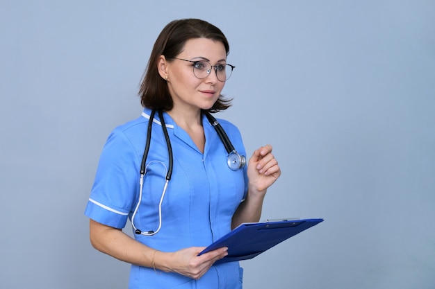 Retrato de mujer madura enfermera con portapapeles. Mujer en uniforme azul con estetoscopio sobre fondo gris, espacio de copia