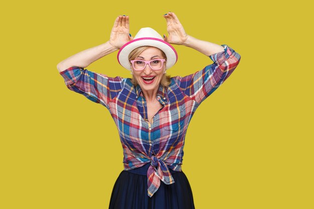 Foto retrato de mujer madura con estilo moderno divertido en estilo casual con sombrero y anteojos de pie con gesto de orejas de conejo y mirando con gran sonrisa. tiro del estudio de interior aislado en fondo amarillo.