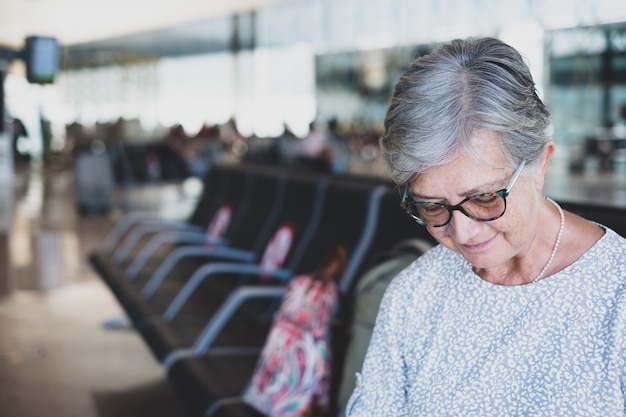 Retrato de mujer madura sentada en el aeropuerto con equipaje mediante teléfono móvil esperando la salida del vuelo. Coronavirus y concepto de libertad.