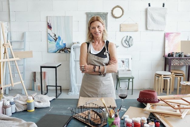 Retrato de mujer madura sonriente en delantal de pie con los brazos cruzados en el estudio de arte con imágenes en la pared