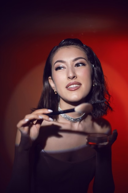 Retrato de una mujer maquilladora de moda sexy con una falda blanca con un cepillo de polvo negro y soportes de caja en el fondo de luz roja del estudio