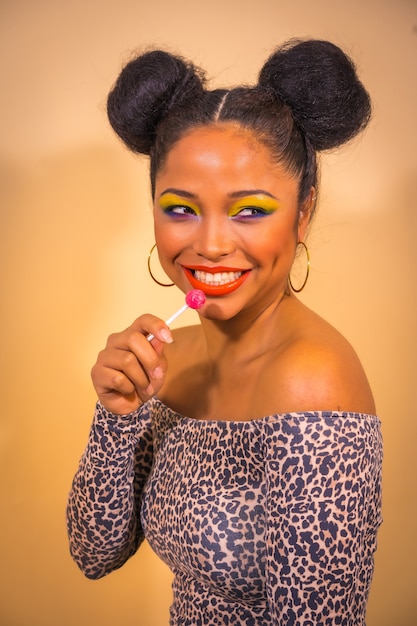 Retrato de una mujer con maquillaje brillante y peinado de moño de dos lados sosteniendo una piruleta