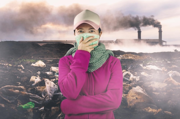 Foto retrato de una mujer con una máscara de contaminación mientras está de pie frente a una fábrica que emite humo