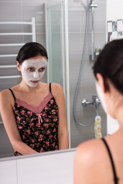 Retrato de mujer con máscara cosmética en la cara mirando a sí misma en el espejo en el moderno baño de azulejos en casa
