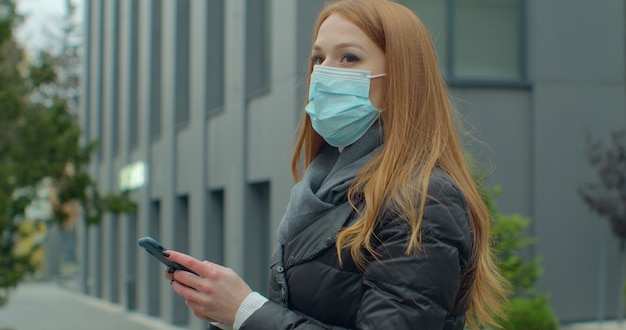 Retrato de mujer con máscara protectora médica escribiendo por teléfono móvil al aire libre.