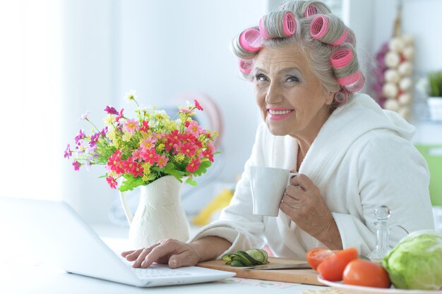 Retrato de mujer mayor en albornoz con rulos sentado en la mesa con portátil