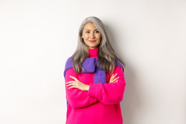 Retrato de mujer mayor asiática confiada con los brazos cruzados en el pecho y sonriendo, de pie sobre fondo blanco en ropa de moda