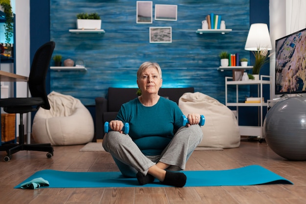 Retrato de mujer mayor atleta mirando a la cámara sentado en posición de loto en la estera de yoga en la sala de estar durante el entrenamiento de bienestar