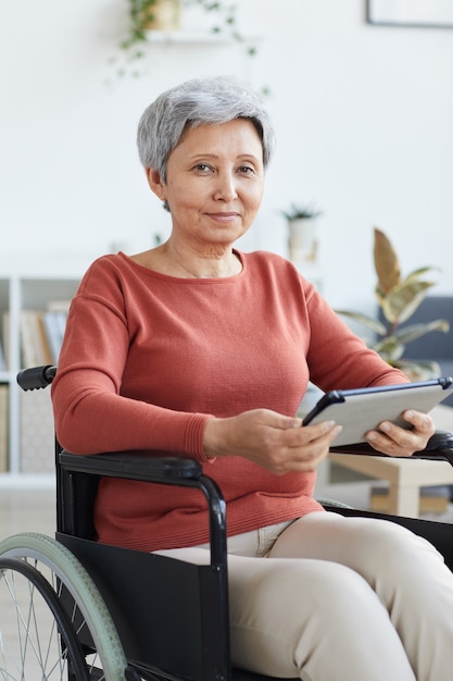 Retrato de mujer mayor discapacitada sentada en silla de ruedas mientras usa tablet pc en casa