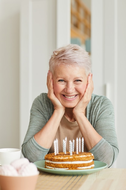 Retrato de mujer mayor feliz sentado a la mesa con pastel con velas y sonriendo a la cámara
