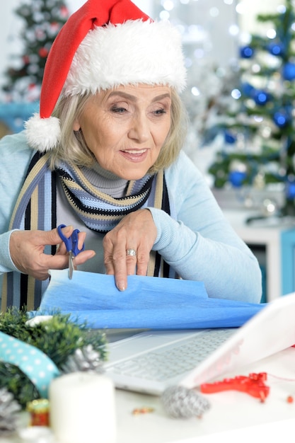 Retrato de mujer mayor con gorro de Papá Noel sentado en la mesa con el portátil y preparándose para la Navidad