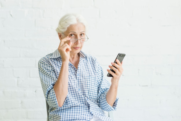 Foto retrato de una mujer mayor que sostiene teléfono inteligente frente a la pared blanca