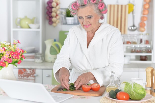Retrato de mujer mayor en rulos cortando verduras en la mesa de la cocina