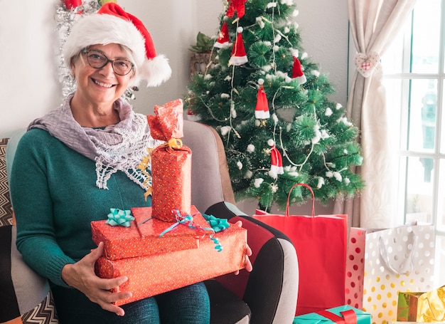 Retrato de una mujer mayor sonriente con un sombrero de Santa Claus sosteniendo muchos regalos de Navidad para la familia - Feliz Navidad en casa para un anciano jubilado disfrutando de las vacaciones