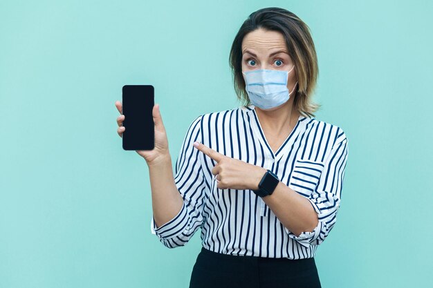 Retrato de mujer de mediana edad conmocionada con mascarilla médica quirúrgica de pie y mostrando móvil inteligente