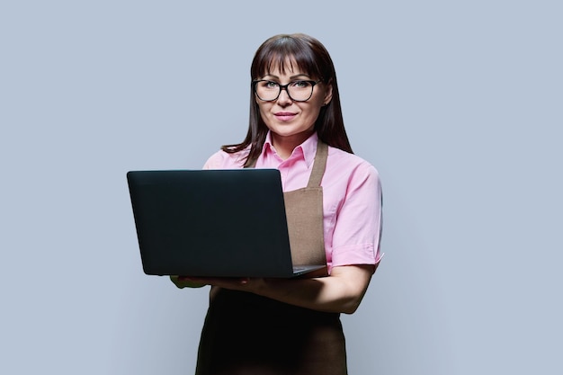 Retrato de una mujer de mediana edad con un delantal usando una computadora portátil sobre un fondo gris