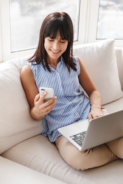 Retrato de mujer de mediana edad en ropa elegante con teléfono celular y computadora portátil mientras está sentado en el sofá en apartamento luminoso